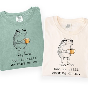 May include: Two t-shirts, one bay green and one ivory, each featuring a cartoon frog holding a mug. The frog has a weary expression. Below the frog is the text "God is still working on me."