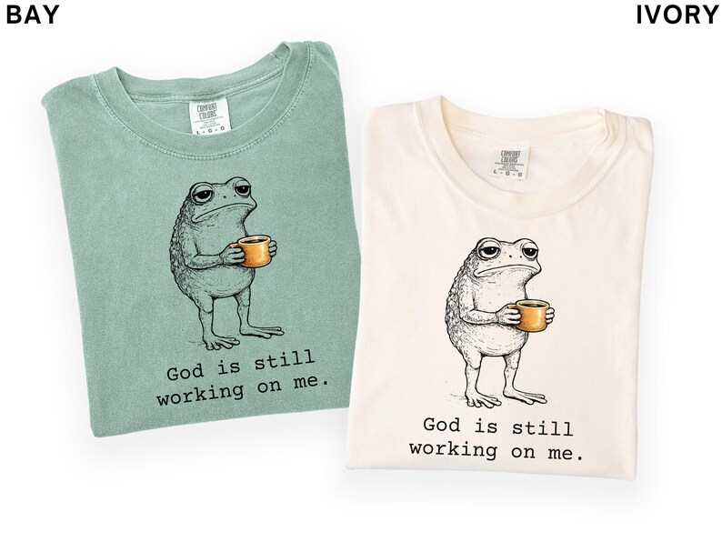 May include: Two t-shirts, one bay green and one ivory, each featuring a cartoon frog holding a mug. The frog has a weary expression. Below the frog is the text "God is still working on me."