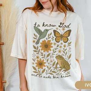 May include: Ivory-colored t-shirt featuring a nature-themed graphic. The design includes a hummingbird, butterfly, flowers, and a frog, with the text "to know God and to make Him known".