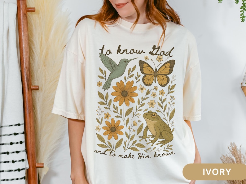 May include: Ivory-colored t-shirt featuring a nature-themed graphic. The design includes a hummingbird, butterfly, flowers, and a frog, with the text "to know God and to make Him known".