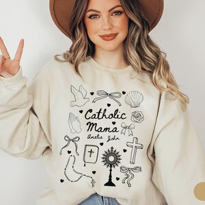 May include: A beige sweatshirt with a graphic design of religious symbols and the text "Catholic Mama Amelia John".