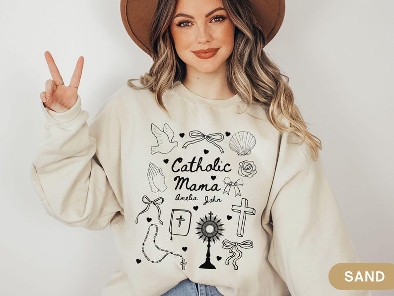 May include: A beige sweatshirt with a graphic design of religious symbols and the text "Catholic Mama Amelia John".