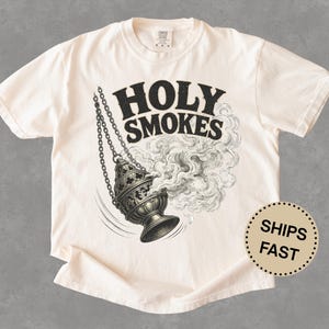May include: Cream-colored t-shirt featuring the text "HOLY SMOKES" above a detailed black and white illustration of a censer with smoke billowing out. The shirt has short sleeves and a crew neck.