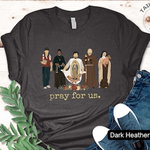 May include: Dark heather grey t-shirt with a graphic of five figures standing in front of a floral design. The text "pray for us." is printed below the graphic.