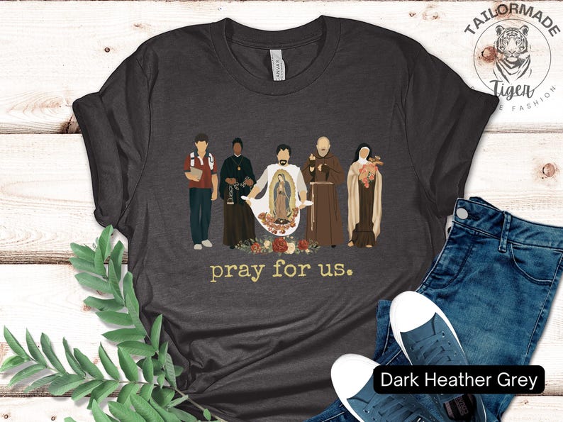May include: Dark heather grey t-shirt with a graphic of five figures standing in front of a floral design. The text "pray for us." is printed below the graphic.