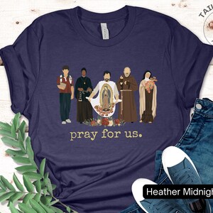 May include: A navy blue t-shirt with a graphic of five figures standing in front of a floral design. The text "pray for us." is printed below the graphic.