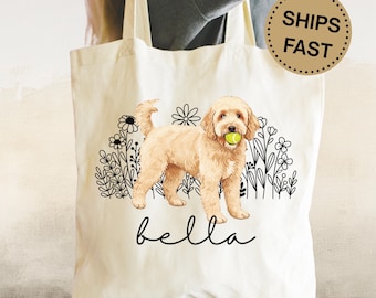 May include: Cream tote bag with a golden doodle dog illustration holding a tennis ball, with black floral designs. The name "bella" is written in cursive below the dog. The bag is held by a person and has a simple design.