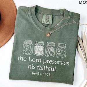Puede incluir: Una camiseta de manga corta color verde musgo con un gr&aacute;fico blanco de cuatro frascos de vidrio llenos de pepinillos, bayas, esp&aacute;rragos y fruta. Debajo de los frascos, el texto "the Lord preserves his faithful. Psalm 31:23" aparece en letras blancas.