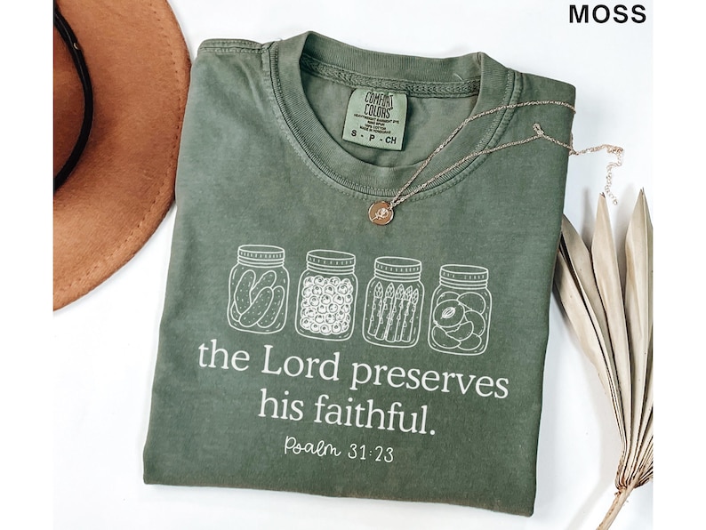 Puede incluir: Una camiseta de manga corta color verde musgo con un gr&aacute;fico blanco de cuatro frascos de vidrio llenos de pepinillos, bayas, esp&aacute;rragos y fruta. Debajo de los frascos, el texto "the Lord preserves his faithful. Psalm 31:23" aparece en letras blancas.