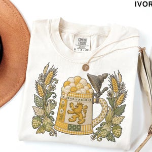 Puede incluir: Una camiseta de color marfil con un gráfico de una jarra de cerveza con un emblema de león, rodeada de tallos de trigo y lúpulo. La camiseta tiene una etiqueta de la marca Comfort Colors en el cuello con la talla S y el texto IVORY arriba.