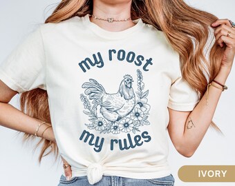 Könnte beinhalten: Ein weißes T-Shirt mit dem Text "my roost my rules" in blauer Farbe bedruckt. Eine blaue Illustration eines Huhns mit Blumen ist mittig auf dem T-Shirt platziert.