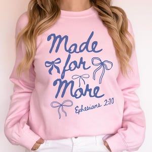 Puede incluir: Sudadera rosa claro con la frase "Made for More" en azul, adornada con dibujos de lazos. Tambi&eacute;n incluye la referencia b&iacute;blica "Efesios 2:10". Es una sudadera de manga larga y cuello redondo.