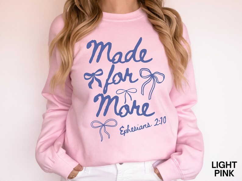 Puede incluir: Sudadera rosa claro con la frase "Made for More" en azul, adornada con dibujos de lazos. Tambi&eacute;n incluye la referencia b&iacute;blica "Efesios 2:10". Es una sudadera de manga larga y cuello redondo.