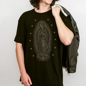Puede incluir: Camiseta negra con una ilustraci&oacute;n detallada de Nuestra Se&ntilde;ora de Guadalupe, rodeada de estrellas. El gr&aacute;fico es en tonos grises y marrones. La persona lleva una chaqueta negra sobre el hombro.