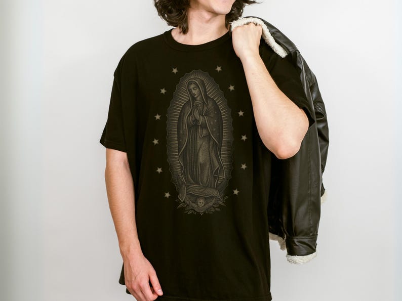 Puede incluir: Camiseta negra con una ilustraci&oacute;n detallada de Nuestra Se&ntilde;ora de Guadalupe, rodeada de estrellas. El gr&aacute;fico es en tonos grises y marrones. La persona lleva una chaqueta negra sobre el hombro.