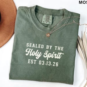 May include: Moss green t-shirt with the text "SEALED BY THE Holy Spirit EST 03.13.26" in white. A gold-colored necklace with a pendant is draped on the shirt. A brown hat is partially visible in the upper left corner.
