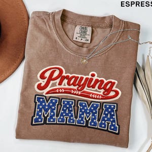 Puede incluir: Camiseta color espresso con las palabras "Praying MAMA" bordadas en rojo y azul. La camiseta tiene cuello redondo y una pequeña etiqueta en la parte superior del pecho. Un collar plateado con un colgante está sobre la camiseta.