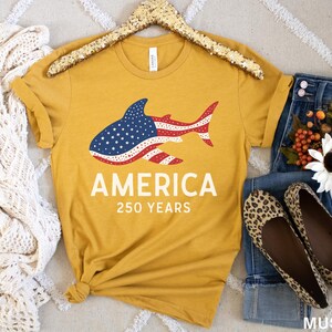 Puede incluir: Camiseta amarilla mostaza con un dise&ntilde;o de tibur&oacute;n con los colores de la bandera americana. La palabra "AMERICA" y "250 YEARS" est&aacute;n impresas debajo. La camiseta se combina con vaqueros, bailarinas con estampado de leopardo y una bufanda blanca.