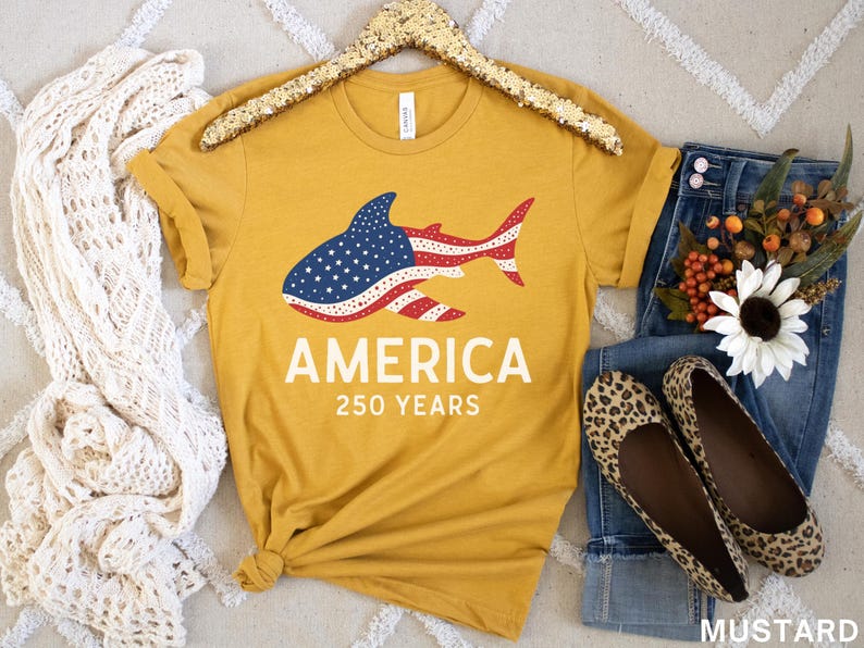 Puede incluir: Camiseta amarilla mostaza con un dise&ntilde;o de tibur&oacute;n con los colores de la bandera americana. La palabra "AMERICA" y "250 YEARS" est&aacute;n impresas debajo. La camiseta se combina con vaqueros, bailarinas con estampado de leopardo y una bufanda blanca.