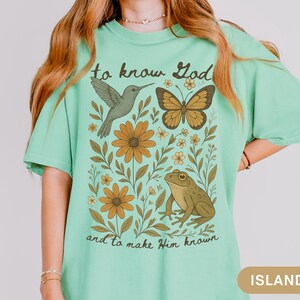 May include: A light green t-shirt with a nature-themed graphic. The design includes a hummingbird, butterfly, frog, and flowers. The text "to know God and to make Him known" is printed on the shirt.