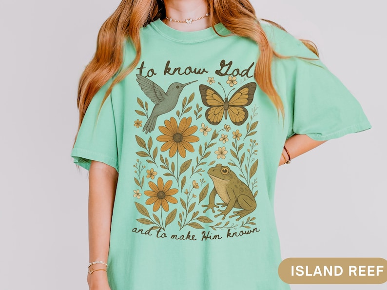 May include: A light green t-shirt with a nature-themed graphic. The design includes a hummingbird, butterfly, frog, and flowers. The text "to know God and to make Him known" is printed on the shirt.