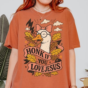 Puede incluir: Camiseta de color &oacute;xido con un dibujo de un ganso de dibujos animados con gafas y un lazo, rodeado de hojas y nubes. El texto "HONK IF YOU LOVE JESUS" aparece en una pancarta. La camiseta es de estilo c&oacute;modo e informal.