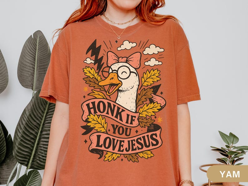 Puede incluir: Camiseta de color &oacute;xido con un dibujo de un ganso de dibujos animados con gafas y un lazo, rodeado de hojas y nubes. El texto "HONK IF YOU LOVE JESUS" aparece en una pancarta. La camiseta es de estilo c&oacute;modo e informal.
