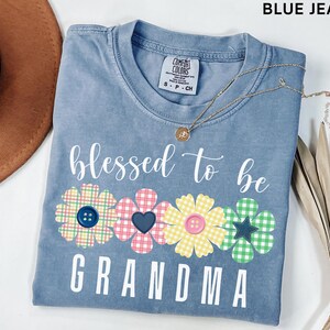以下が含まれることがあります： 「blessed to be grandma」の白文字がプリントされた、ブルーデニムカラーのTシャツ。ボタンが中央にある3つの花の模様が特徴です。金色のネックレスとペンダントがシャツの上に置かれています。