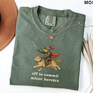 May include: Moss green Comfort Colors t-shirt with a humorous illustration of a frog knight on a cat, holding a sword. The text "off to commit minor heroics" is printed below. A silver necklace and a brown hat are also visible in the image.