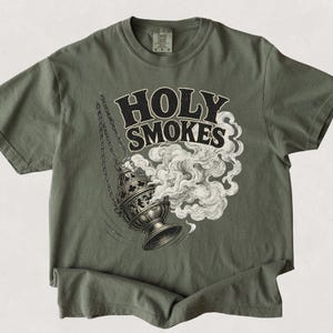 May include: A moss green t-shirt featuring the words "HOLY SMOKES" in a bold, vintage font. Below the text is a detailed illustration of a censer with smoke billowing out. The shirt is made of a soft material.