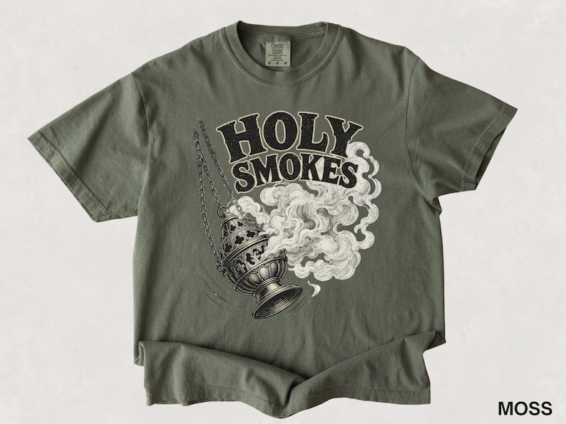 May include: A moss green t-shirt featuring the words "HOLY SMOKES" in a bold, vintage font. Below the text is a detailed illustration of a censer with smoke billowing out. The shirt is made of a soft material.
