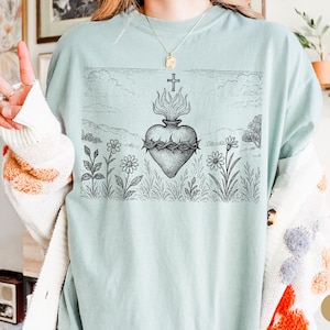 K&ouml;nnte beinhalten: Hellblaues T-Shirt mit einer schwarz-wei&szlig;en Grafik eines Herzens mit Flammen, einer Dornenkrone und einem Kreuz. Das Design ist vor einer Landschaft mit Blumen und Himmel platziert. Das T-Shirt hat eine lockere Passform.