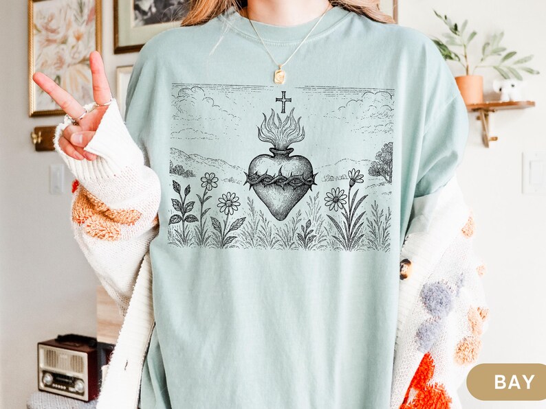 K&ouml;nnte beinhalten: Hellblaues T-Shirt mit einer schwarz-wei&szlig;en Grafik eines Herzens mit Flammen, einer Dornenkrone und einem Kreuz. Das Design ist vor einer Landschaft mit Blumen und Himmel platziert. Das T-Shirt hat eine lockere Passform.