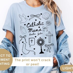 May include: Light blue t-shirt with a graphic design of religious symbols, including a dove, a cross, a rosary, and the text "Catholic Mama" with the names "Amelia" and "John".