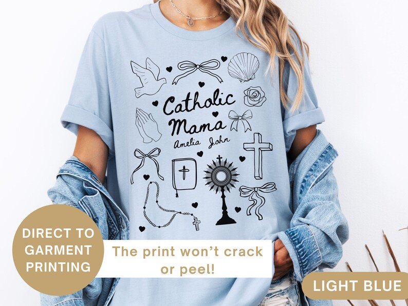 May include: Light blue t-shirt with a graphic design of religious symbols, including a dove, a cross, a rosary, and the text "Catholic Mama" with the names "Amelia" and "John".