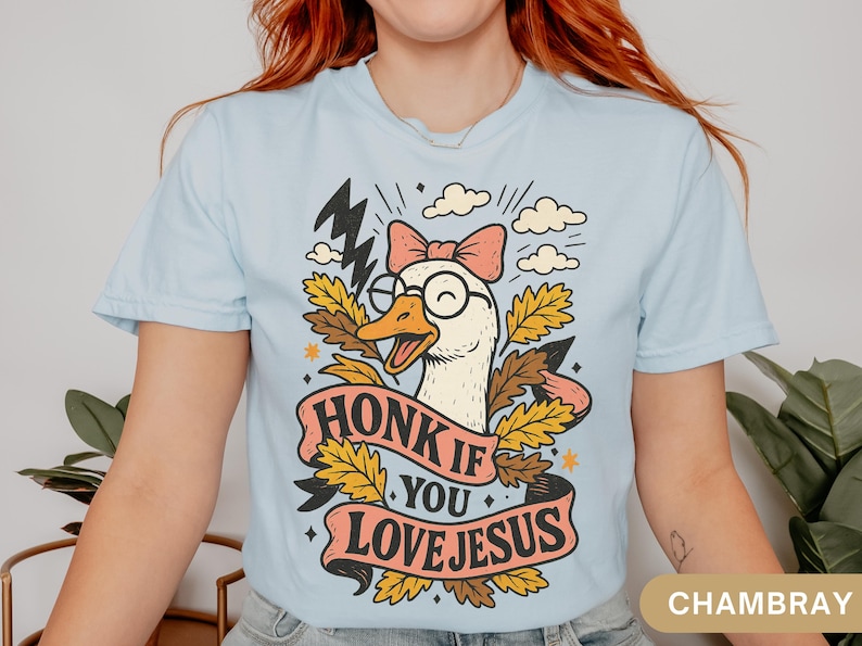 Puede incluir: Camiseta azul claro con un dibujo de un ganso con gafas y un lazo rosa. El ganso est&aacute; rodeado de hojas y el texto "HONK IF YOU LOVE JESUS" aparece en una pancarta. La camiseta es de un material suave.