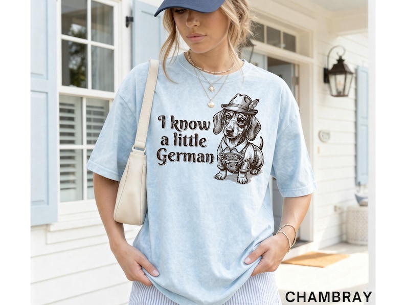 Puede incluir: Una camiseta azul claro con un gr&aacute;fico marr&oacute;n oscuro de un perro salchicha que lleva un sombrero tradicional y lederhosen. El texto "I know a little German" est&aacute; impreso en una fuente marr&oacute;n oscuro junto a la ilustraci&oacute;n del perro.