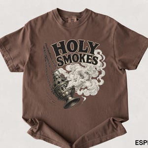 May include: Espresso-colored t-shirt featuring a graphic of a censer with smoke and the words "HOLY SMOKES" in a stylized font. The design is in black and white, contrasting with the shirt's solid color. The shirt is a short-sleeved crew neck.