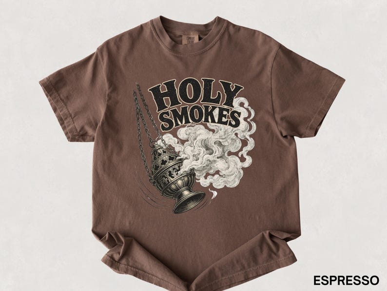 May include: Espresso-colored t-shirt featuring a graphic of a censer with smoke and the words "HOLY SMOKES" in a stylized font. The design is in black and white, contrasting with the shirt's solid color. The shirt is a short-sleeved crew neck.