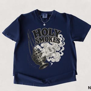 May include: Navy blue t-shirt featuring the text "HOLY SMOKES" above an illustration of a censer with smoke billowing out. The shirt has short sleeves and a round neckline. A silver necklace with a small pendant is visible.