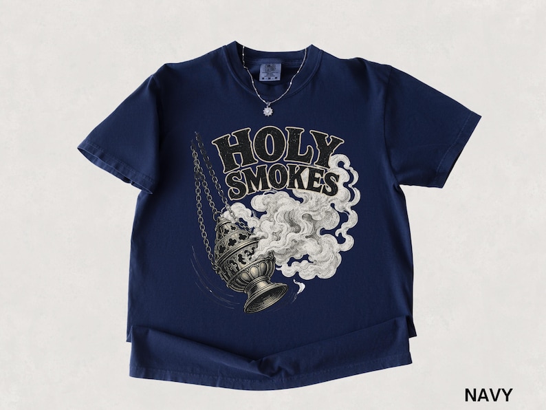 May include: Navy blue t-shirt featuring the text "HOLY SMOKES" above an illustration of a censer with smoke billowing out. The shirt has short sleeves and a round neckline. A silver necklace with a small pendant is visible.