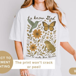 May include: White t-shirt with a nature-themed graphic. The design includes a hummingbird, butterfly, flowers, and a frog, with the text "to know God and to make Him known". The print won't crack or peel.