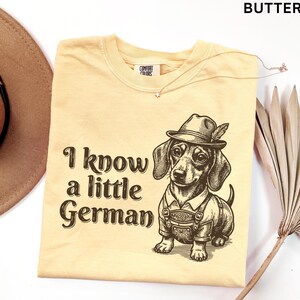Puede incluir: Una camiseta color mantequilla con un gr&aacute;fico marr&oacute;n oscuro de un perro salchicha con un sombrero tradicional y lederhosen. El texto "I know a little German" est&aacute; impreso en una fuente serif marr&oacute;n oscuro a la izquierda del perro.