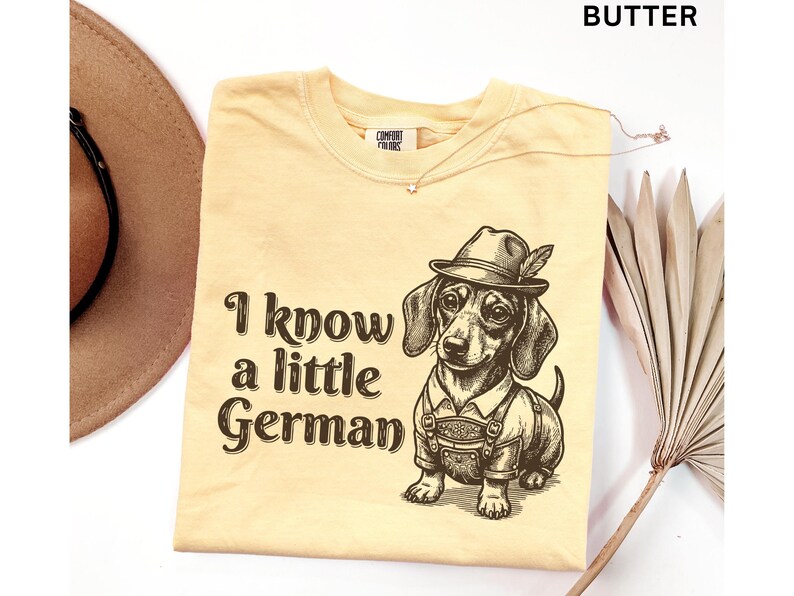 Puede incluir: Una camiseta color mantequilla con un gr&aacute;fico marr&oacute;n oscuro de un perro salchicha con un sombrero tradicional y lederhosen. El texto "I know a little German" est&aacute; impreso en una fuente serif marr&oacute;n oscuro a la izquierda del perro.