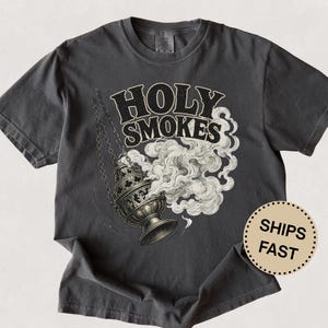 May include: Dark gray t-shirt featuring the words "HOLY SMOKES" in a bold, vintage font above a detailed illustration of a censer with billowing smoke. The design is in black and white.