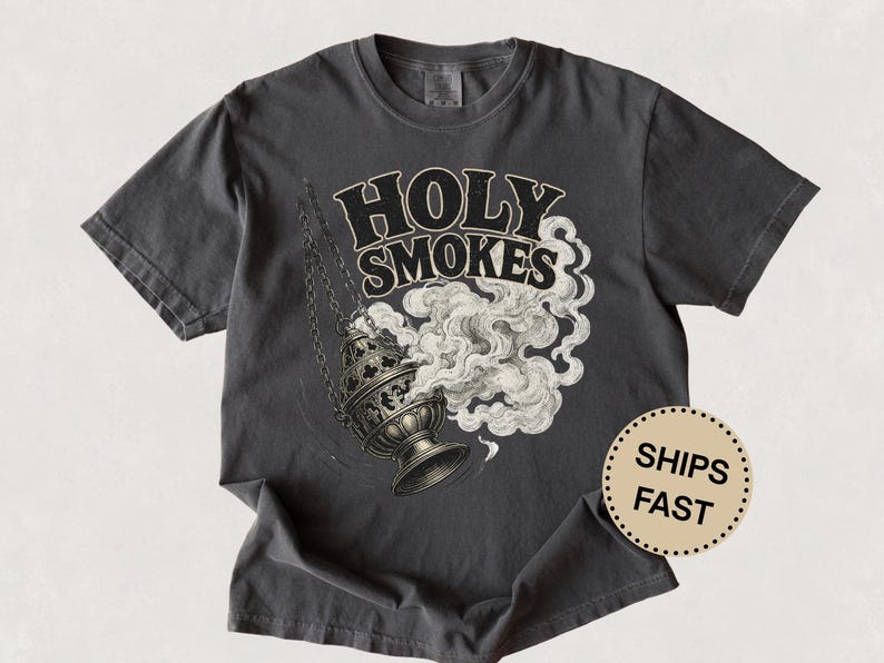 May include: Dark gray t-shirt featuring the words "HOLY SMOKES" in a bold, vintage font above a detailed illustration of a censer with billowing smoke. The design is in black and white.
