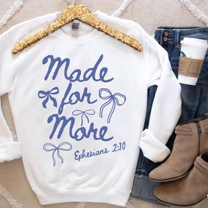 Puede incluir: Sudadera blanca con la frase "Made for More" en azul y dibujos de lazos. La sudadera se muestra con vaqueros azules, botines color canela y una taza de caf&eacute;.