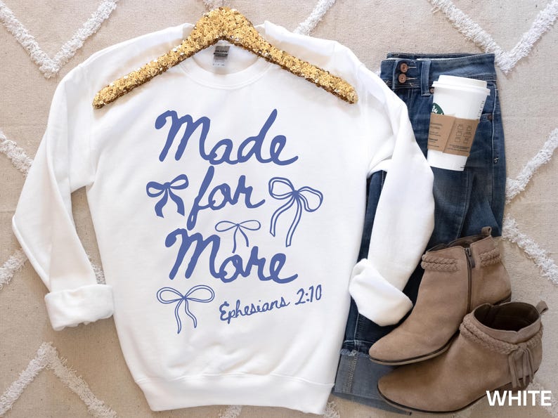 Puede incluir: Sudadera blanca con la frase "Made for More" en azul y dibujos de lazos. La sudadera se muestra con vaqueros azules, botines color canela y una taza de caf&eacute;.