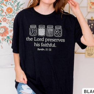 Puede incluir: Una camiseta negra de manga corta con un gr&aacute;fico blanco de cuatro frascos de vidrio llenos de diversos alimentos en conserva. Debajo de los frascos, el texto "the Lord preserves his faithful. Psalm 31:23" aparece en una fuente serif blanca.