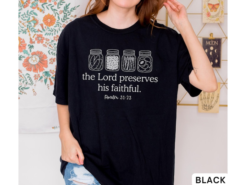 Puede incluir: Una camiseta negra de manga corta con un gr&aacute;fico blanco de cuatro frascos de vidrio llenos de diversos alimentos en conserva. Debajo de los frascos, el texto "the Lord preserves his faithful. Psalm 31:23" aparece en una fuente serif blanca.